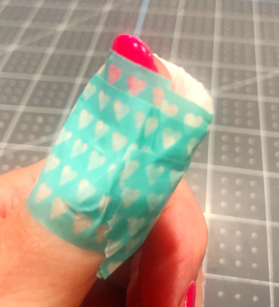 human thumb with a paper napkin on pad of thumb and wrapped in teal washi tape with tiny white hearts as a temporary bandage