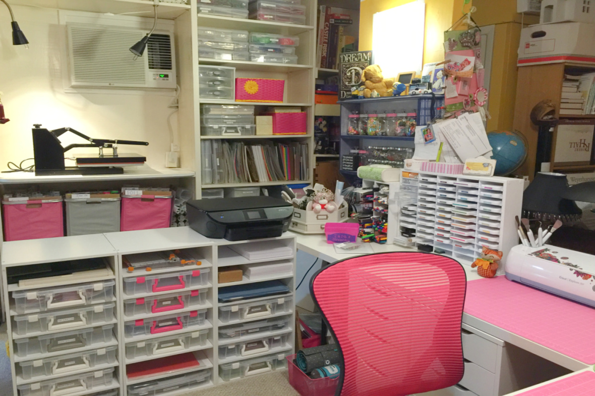 room with white built in bookcase on wall and l shaped desk and row of storage shelves with plastic bins and printer on top and ink storage holder on desk with pink chair at desk for creative space journey