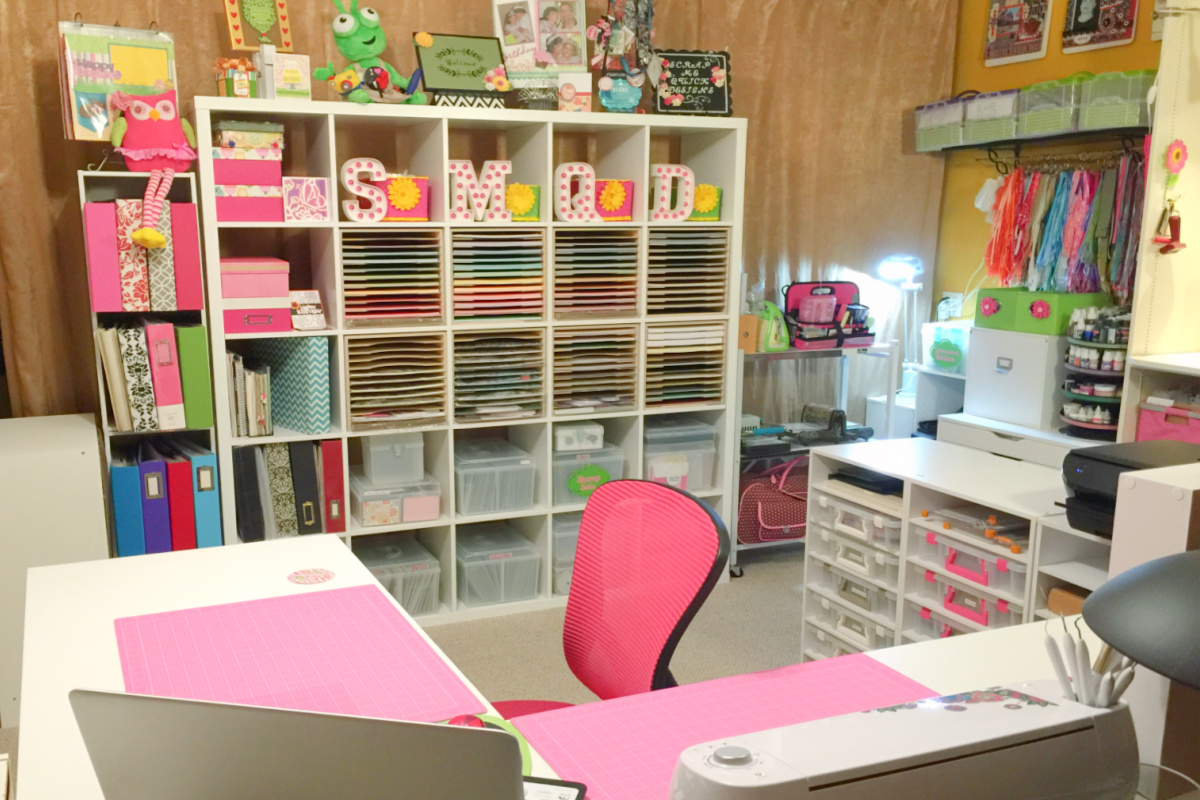 Large white Ikea Bookcase with open shelves filled with scrapbook materials in front of a gold curtain with a white L-shaped desk with a pink chair for my creative space journey